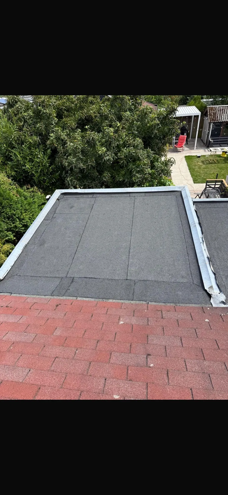 Nieuw bitumen plat dak van bovenaf, met een nette dakrandafwerking en een tuin op de achtergrond